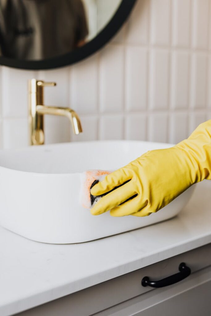 pexels photo 4239110 Crop faceless person in yellow rubber protective glove wiping surface of white sink in bathroom with sponge and cleaning agent